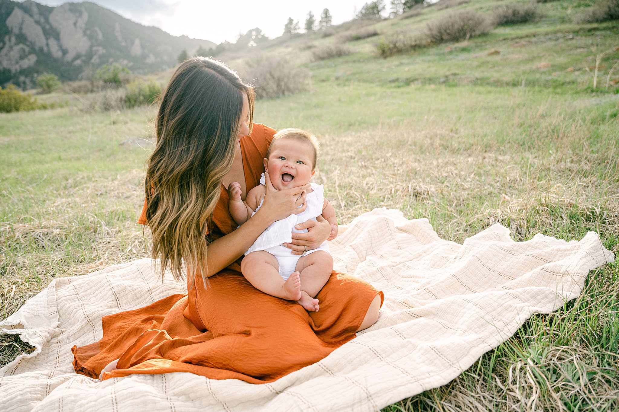 A young mother playing with a blanket with her six month baby during a photo session in Boulder, Colorado with mountains in the background.