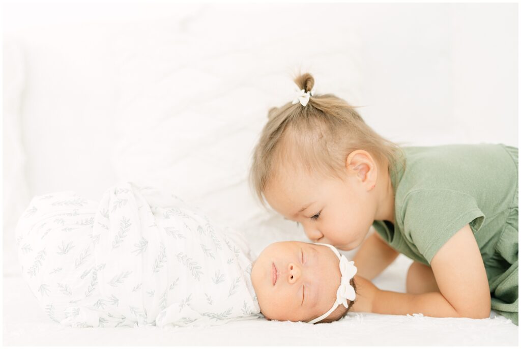 A Colorado toddler kissing her sleeping baby sister during a newborn photoshoot taken by Denver Newborn Photographer Olive and Aster at little white photo studio Frederick Colorado